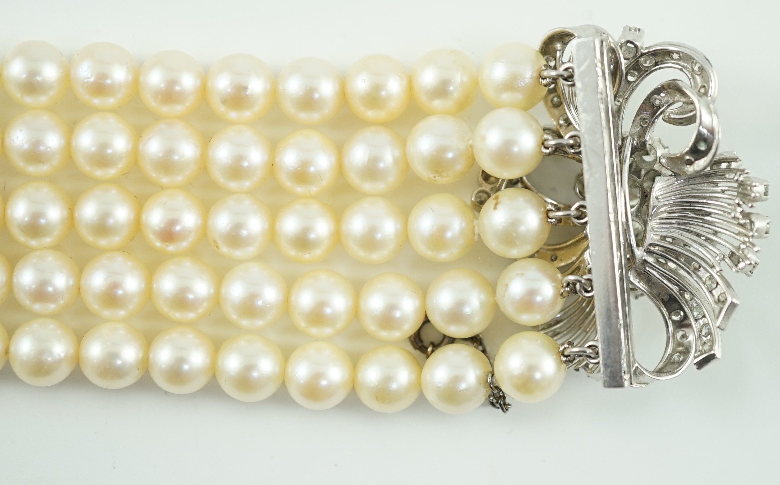 A mid to late 20th century continental quintuple strand cultured pearl bracelet, with white gold and diamond cluster set scroll clasp and diamond set line spacers